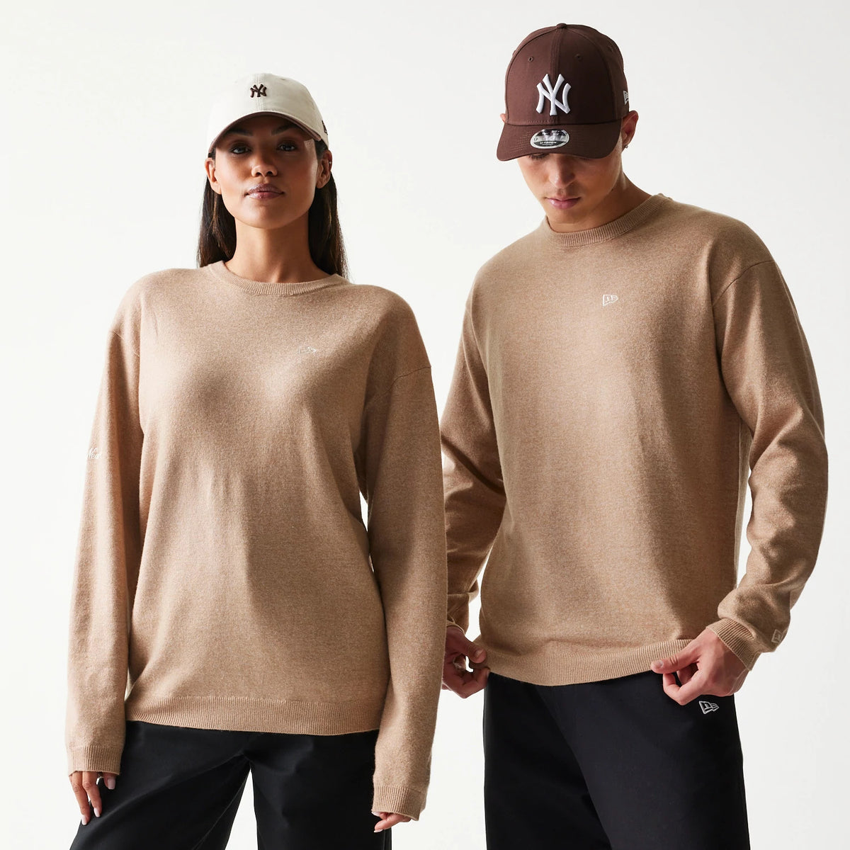 New Era Essential Flag Light Beige Knitted Sweater, on-model front view showing embroidery on the chest and sleeves, with a relaxed fit and ribbed cuffs, styled casually with black trousers and contrasting caps.