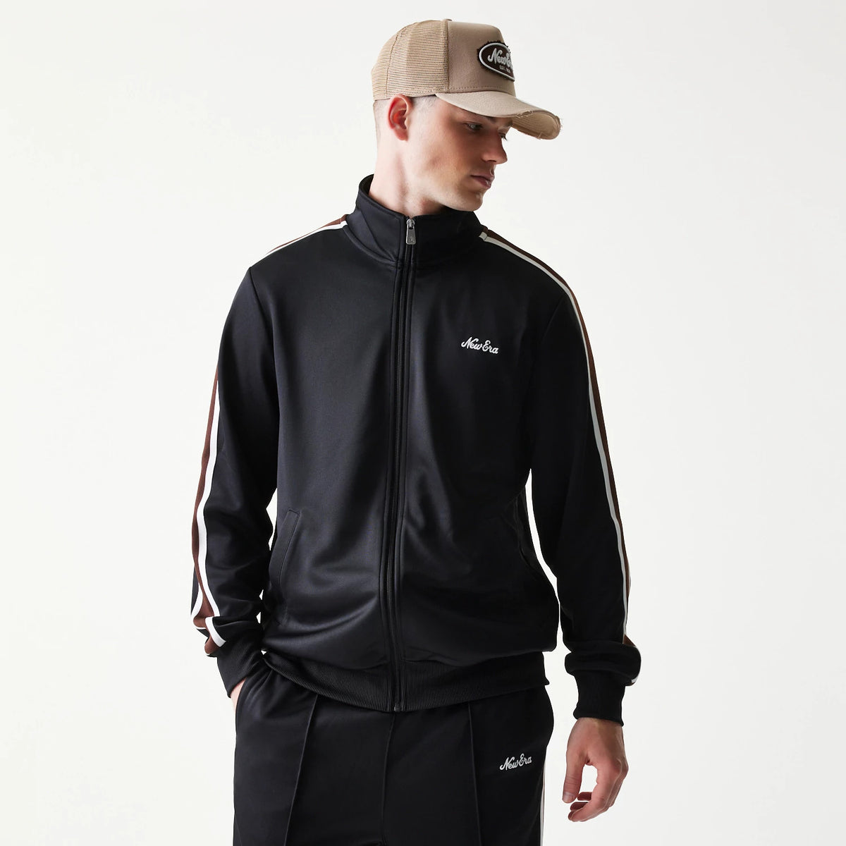 New Era Script Black Track Jacket, front view on a model showing the black jacket with white embroidered script logo on the left chest, zip front, striped sleeves, and matching trousers featuring the same logo on the left leg.