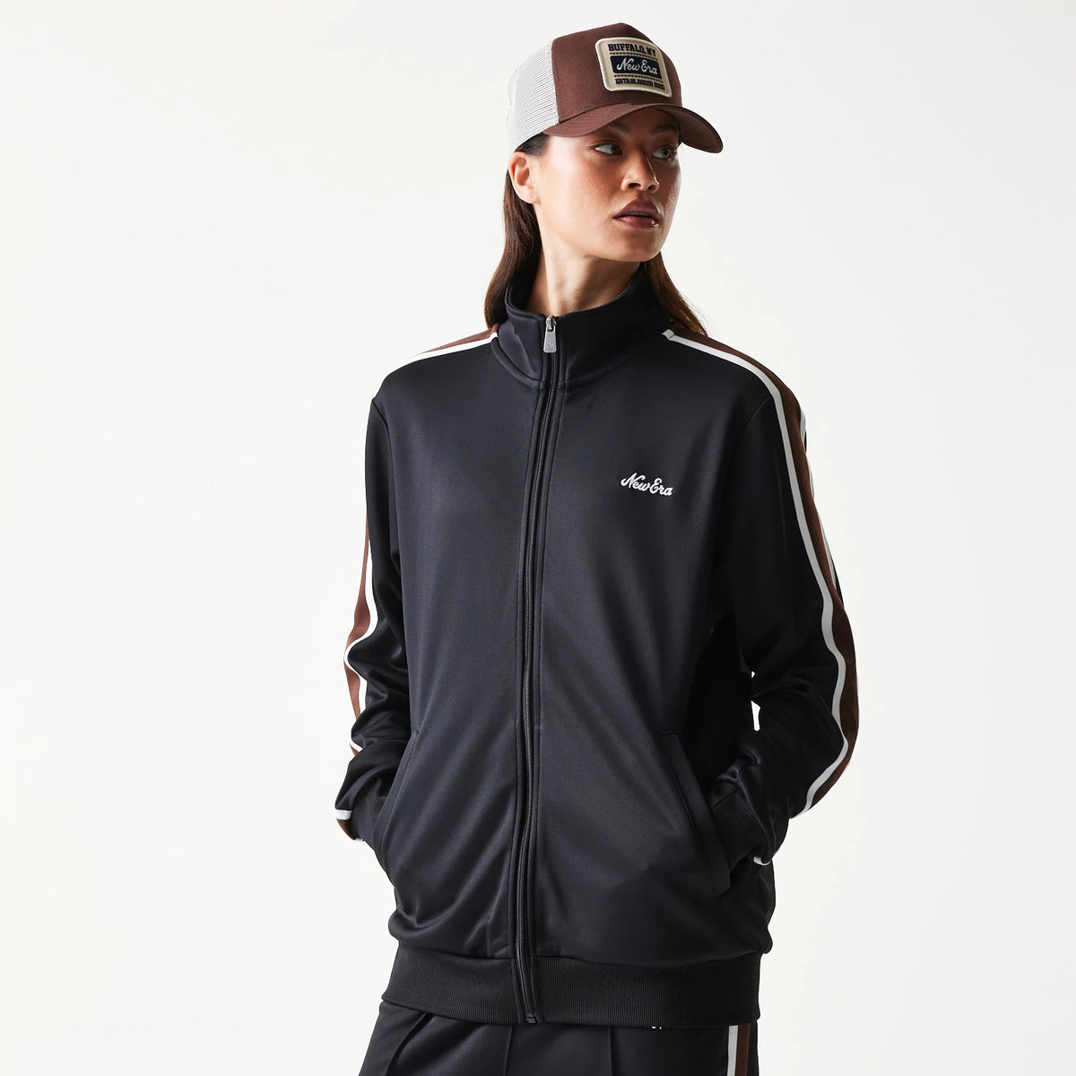 New Era Script Black Track Jacket, front view on a model showing the zipped black jacket with white and brown stripes down the sleeves and an embroidered New Era logo on the left chest, styled with hands in pockets and a matching cap.