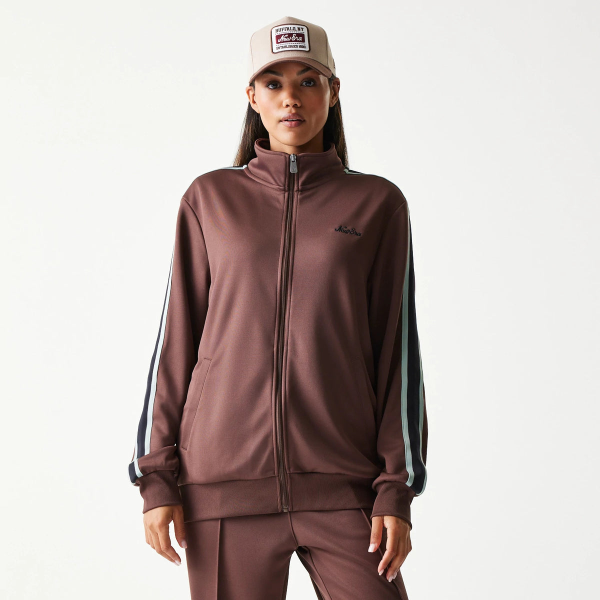 New Era Script Brown Track Jacket, front view showing full zip closure, black embroidered New Era logo on the left chest, and navy and white stripes running down the sleeves, worn by a model with a beige cap.