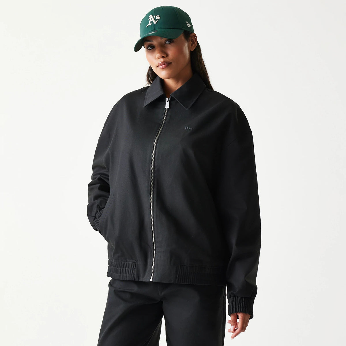 New Era Collared Black Jacket, front view on-model displaying a black design with a front zip closure, an embroidered logo on the chest, side pockets, and elasticated cuffs, worn with matching black trousers and a green cap.