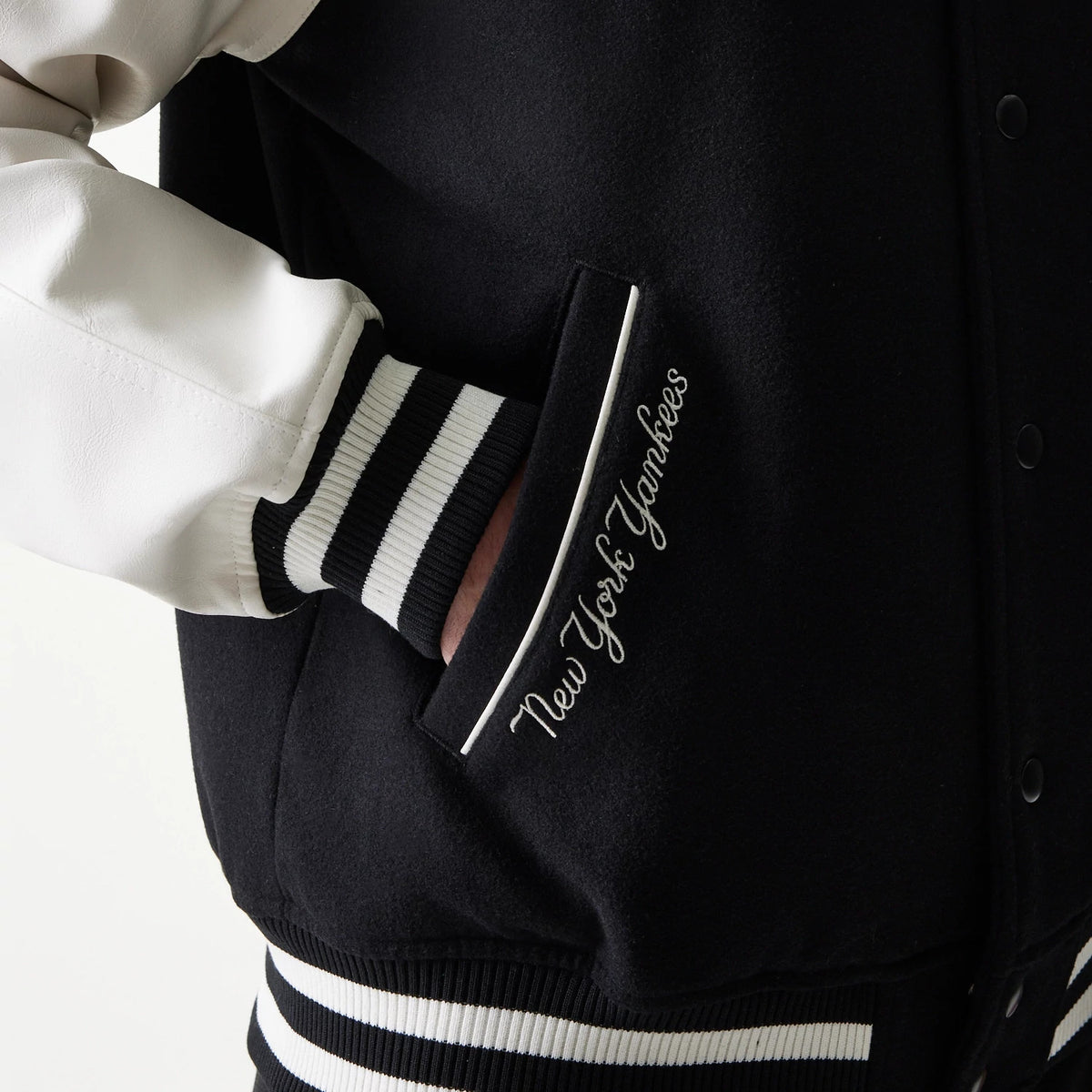 New Era New York Yankees MLB Black Varsity Jacket, side view showing the pocket with white embroidered New York Yankees script and contrasting black and white striped ribbed cuffs on the sleeve and hem.