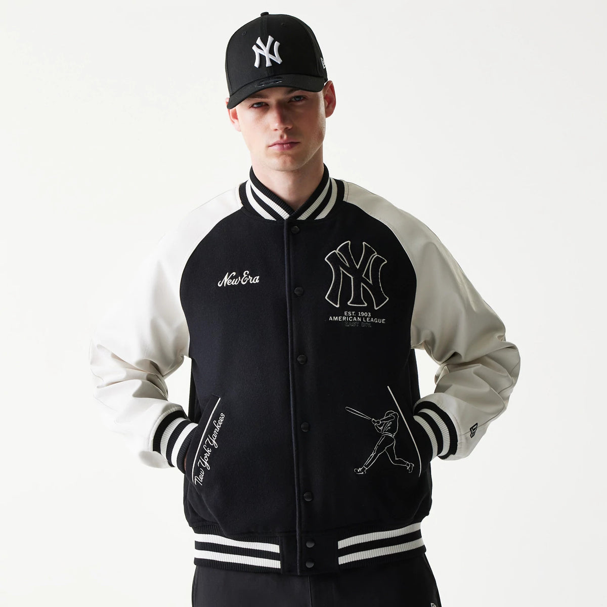 New Era New York Yankees MLB Black Varsity Jacket, front view on-model showing the black body with white sleeves, large embroidered Yankees logo on the chest, New Era script and baseball player graphic near the pocket, and striped collar and cuffs.