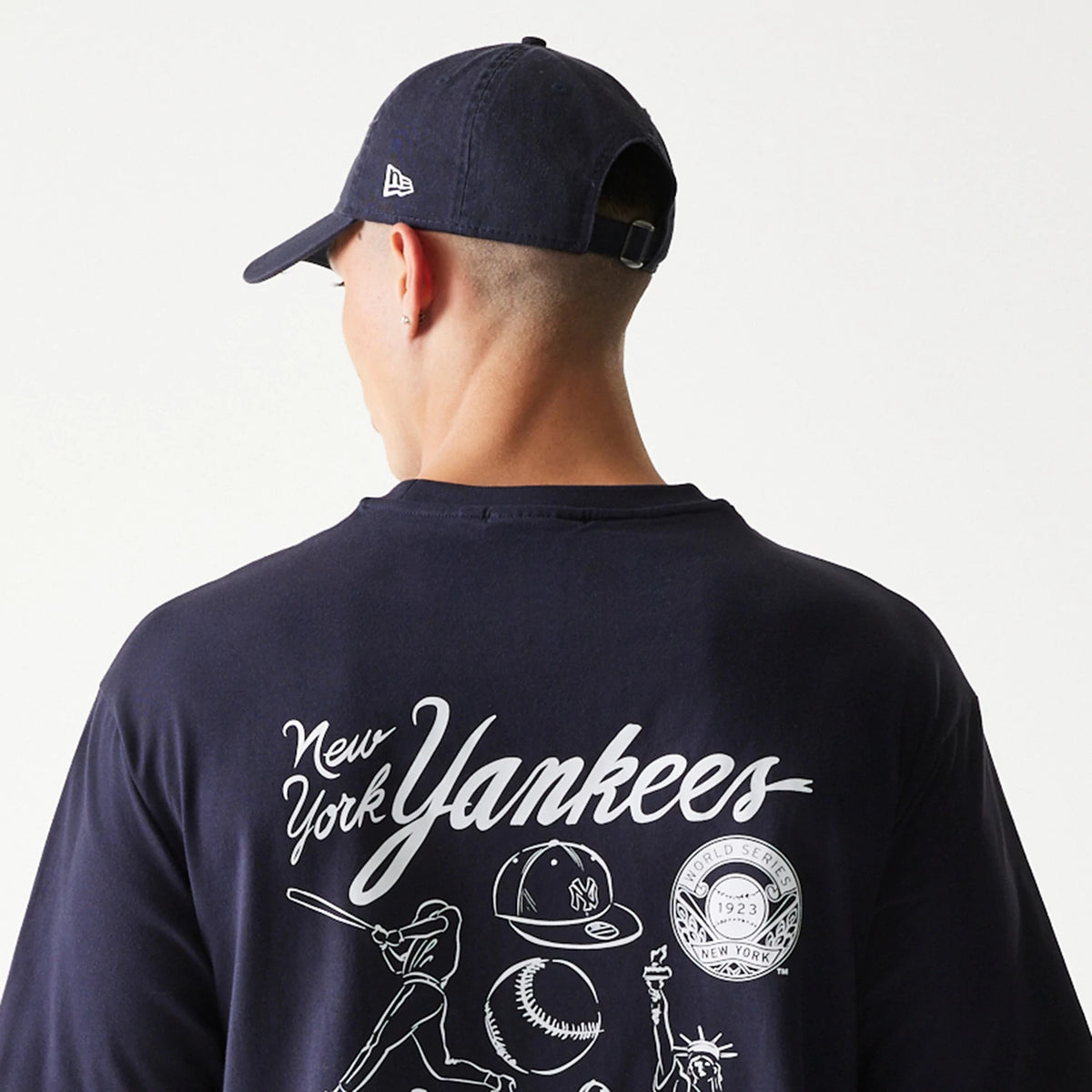 New Era New York Yankees MLB Baseball Graphic Navy Oversized T-Shirt, back view on a model wearing a matching navy cap, featuring large white script New York Yankees lettering and baseball-themed graphics across the upper back.