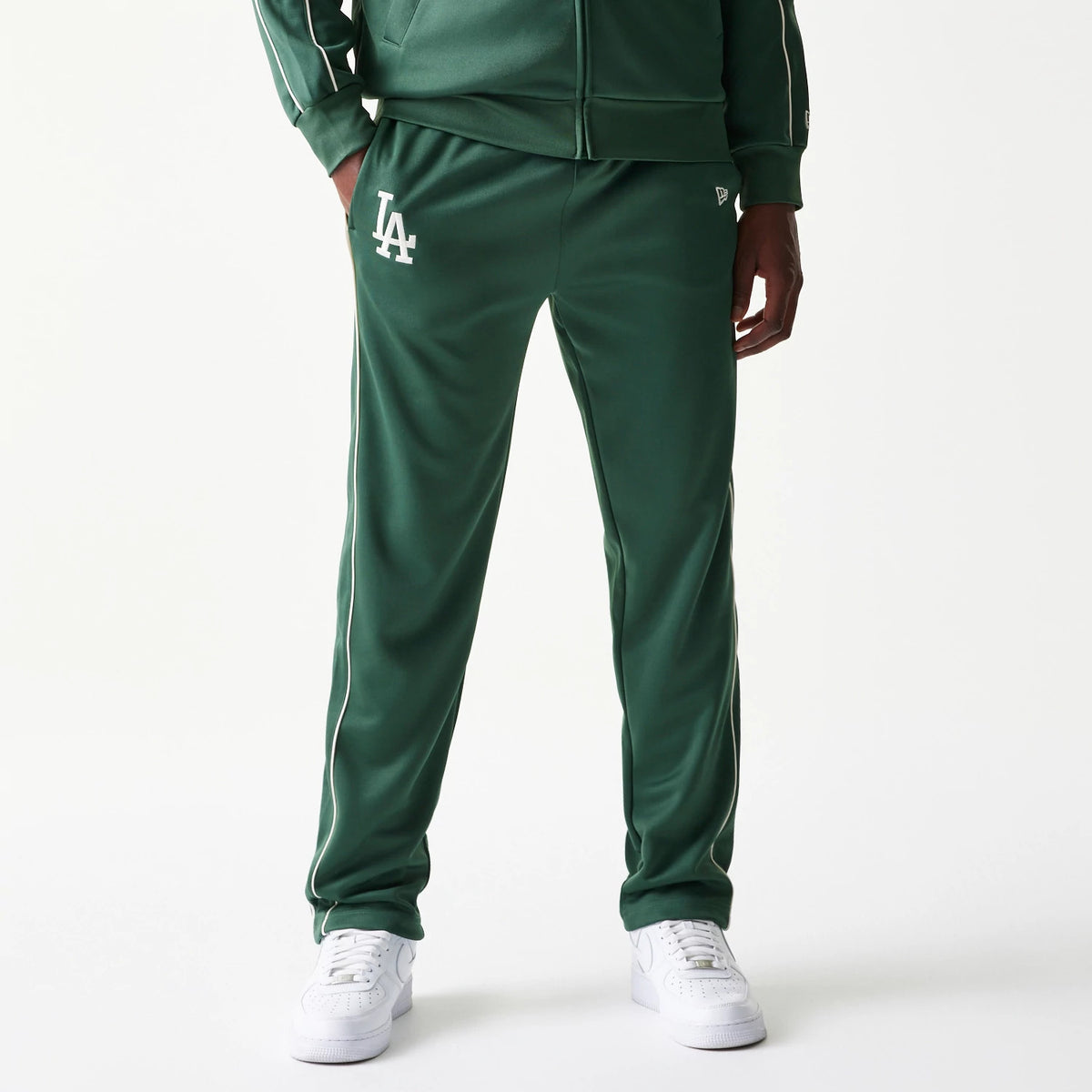 New Era LA Dodgers MLB Piping Dark Green Straight Leg Joggers, front view on a model with white LA logo embroidery on the left thigh, contrasting white piping along the sides, and white trainers.