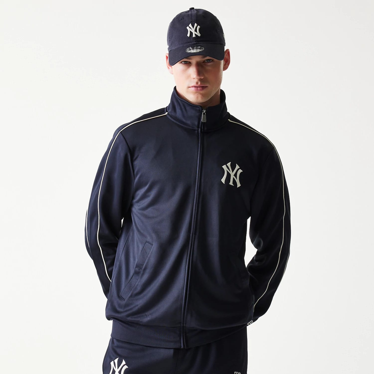 New Era New York Yankees MLB Piping Navy Track Jacket, front view on-model featuring a full zip closure, white embroidered Yankees logo on the left chest, and white piping along the shoulders and sleeves, paired with a matching cap.