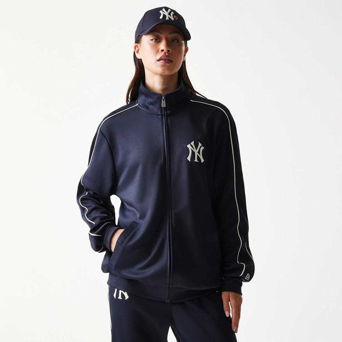 New Era New York Yankees MLB Piping Navy Track Jacket, front view on a model with a matching navy cap, showcasing the New York Yankees logo embroidered on the left chest and white piping along the sleeves and shoulders.