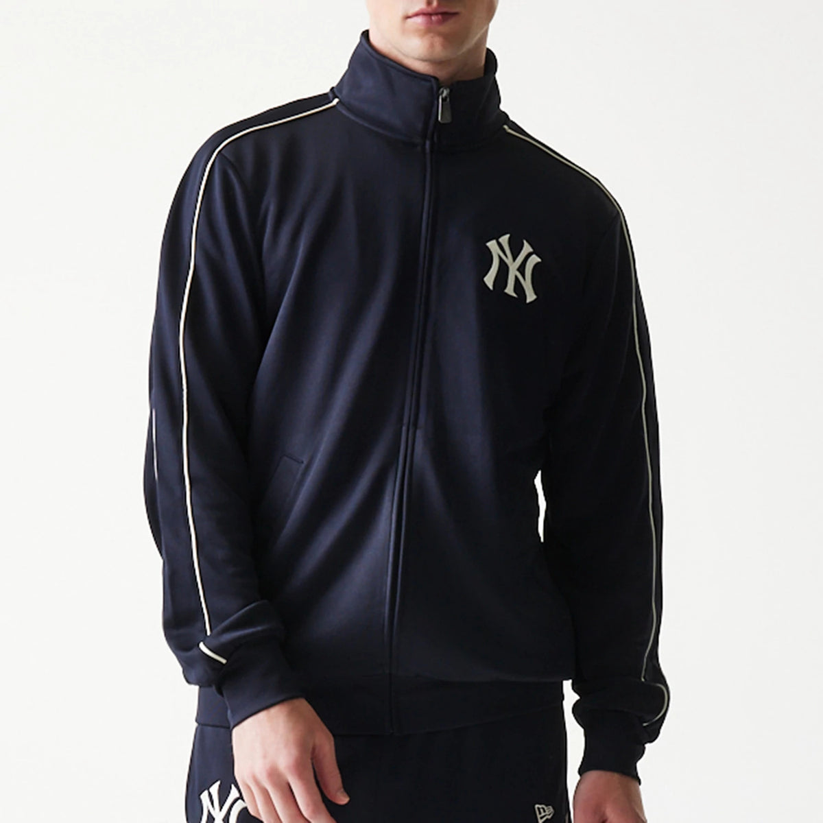 New Era New York Yankees MLB Piping Navy Track Jacket, front view on-model showing the navy jacket with white piping along the shoulders and sleeves, a zipped front, and the white New York Yankees embroidered logo on the left chest.
