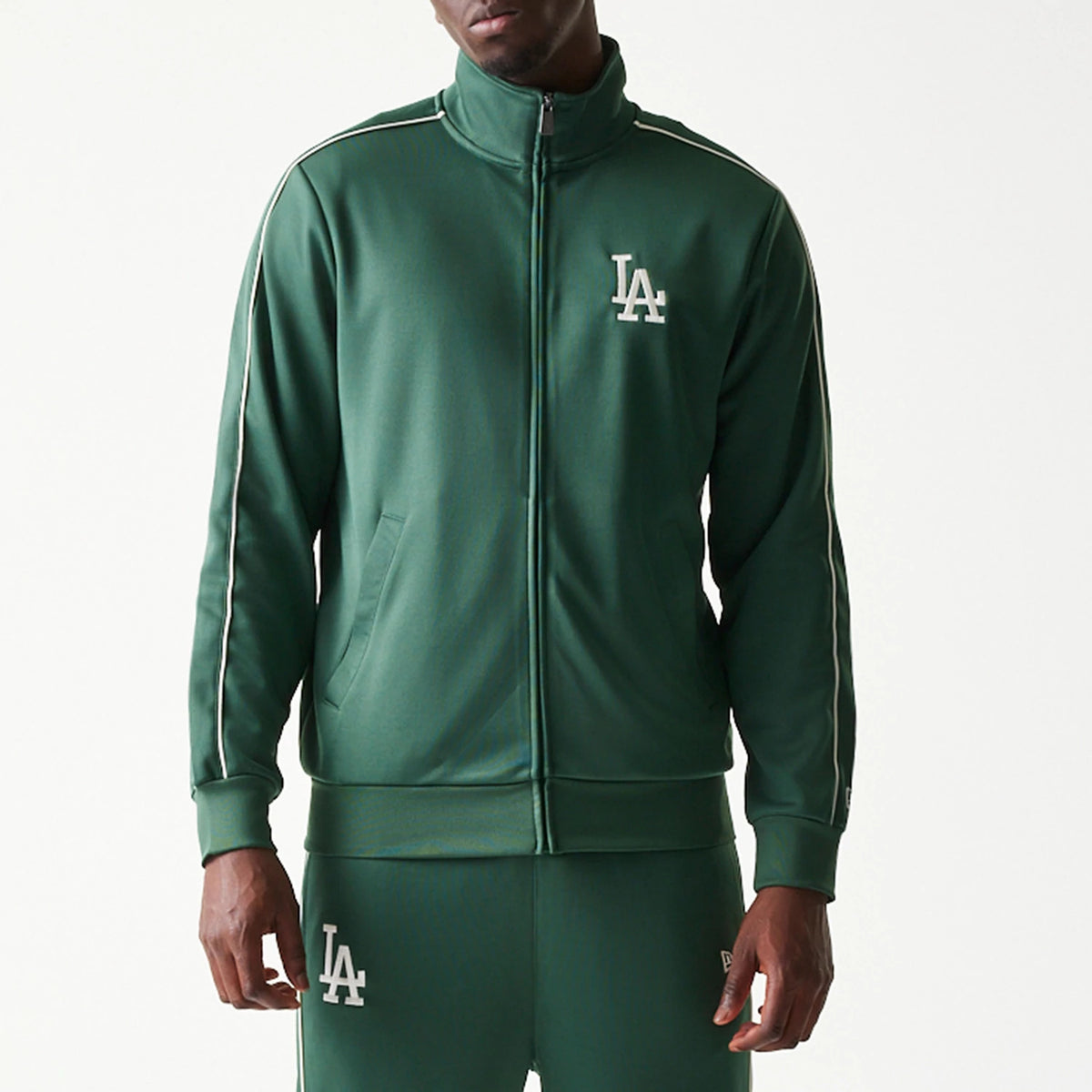 New Era LA Dodgers MLB Piping Dark Green Track Jacket, front view on-model showing the white embroidered LA logo on the left chest, a high collar with a full zip, and white piping detail along the shoulder and sleeves.