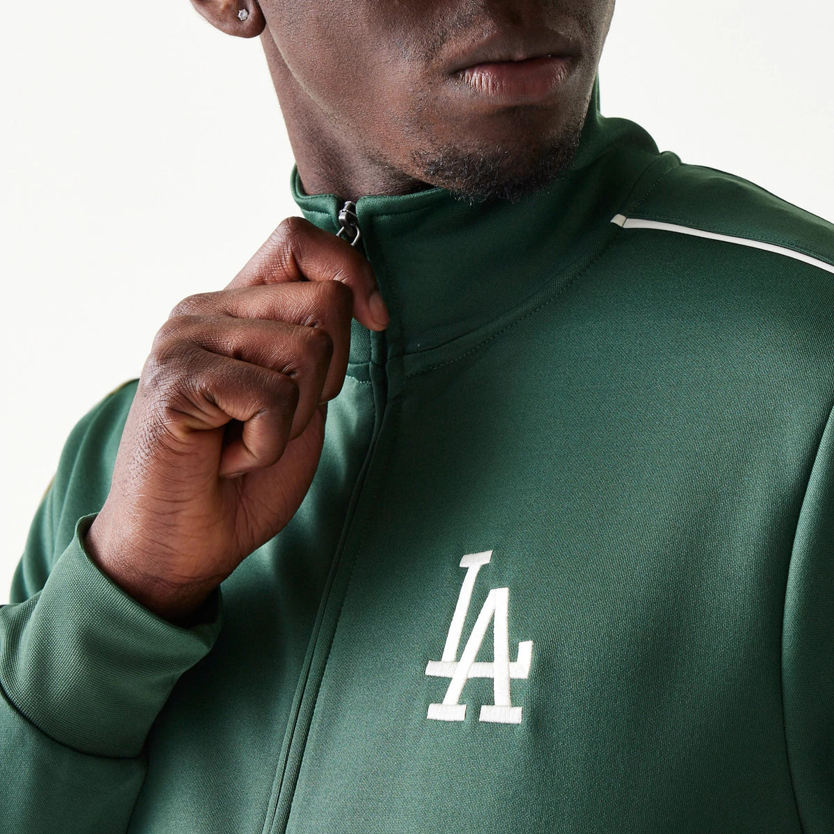 New Era LA Dodgers MLB Piping Dark Green Track Jacket, front view showing the white embroidered LA logo on the chest and a man adjusting the zip, with white piping detail running along the shoulder.