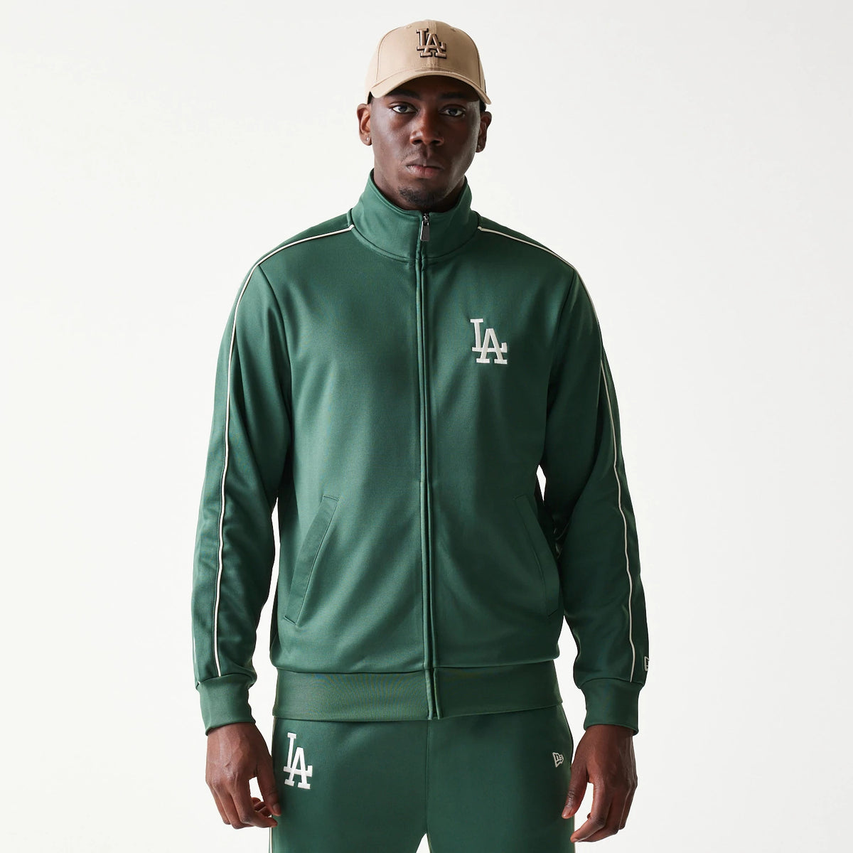 New Era LA Dodgers MLB Piping Dark Green Track Jacket, front view on-model showing the white embroidered LA logo on the left chest, piping details along the sleeves, and a full zip closure, styled with a beige LA cap.