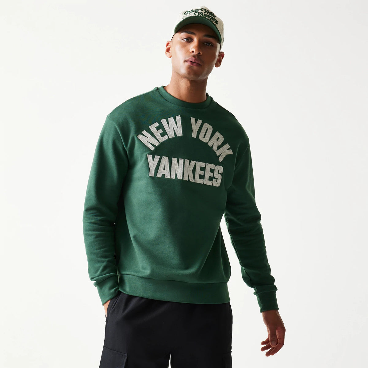 New Era New York Yankees MLB Graphic Dark Green Oversized Crew Neck Sweater, front view on a model wearing a matching cap, featuring large white embroidered New York Yankees lettering across the chest against the dark green fabric.