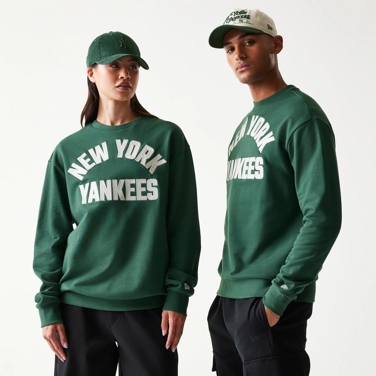 New Era New York Yankees MLB Graphic Dark Green Oversized Crew Neck Sweater, front view on two models showing the large white embroidered New York Yankees lettering across the chest, with one model wearing a matching cap.