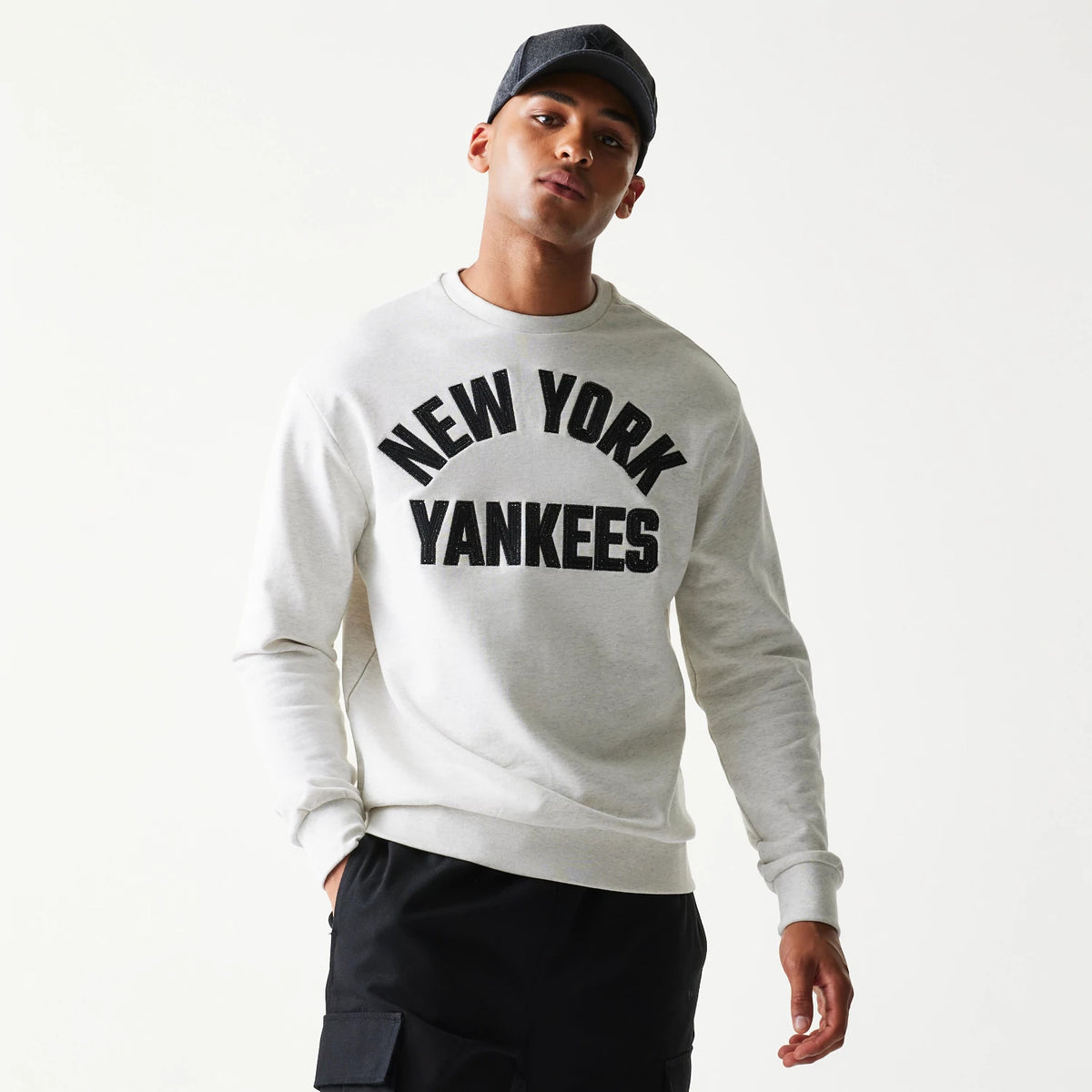 New Era New York Yankees MLB Graphic Pastel Grey Oversized Crew Neck Sweater, front view on a model with a black cap, featuring black 'New York Yankees' lettering across the chest and ribbed cuffs.