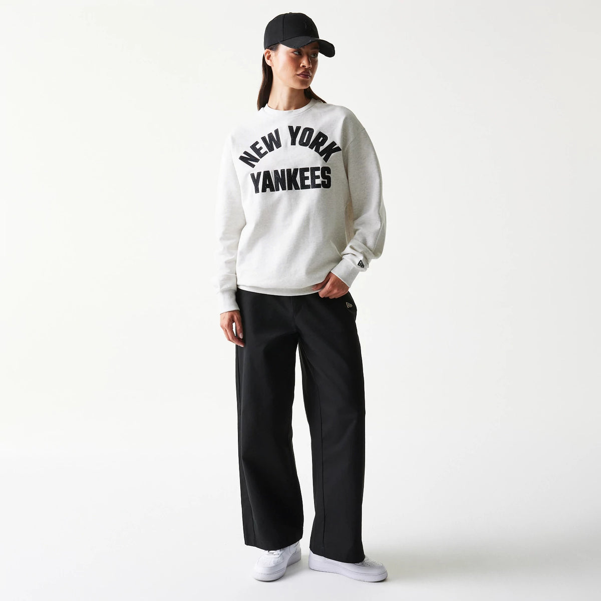 New Era New York Yankees MLB Graphic Pastel Grey Oversized Crew Neck Sweater, on-model view showing large black New York Yankees lettering across the chest, paired with a black cap and trousers, featuring the New Era flag logo on the left sleeve.