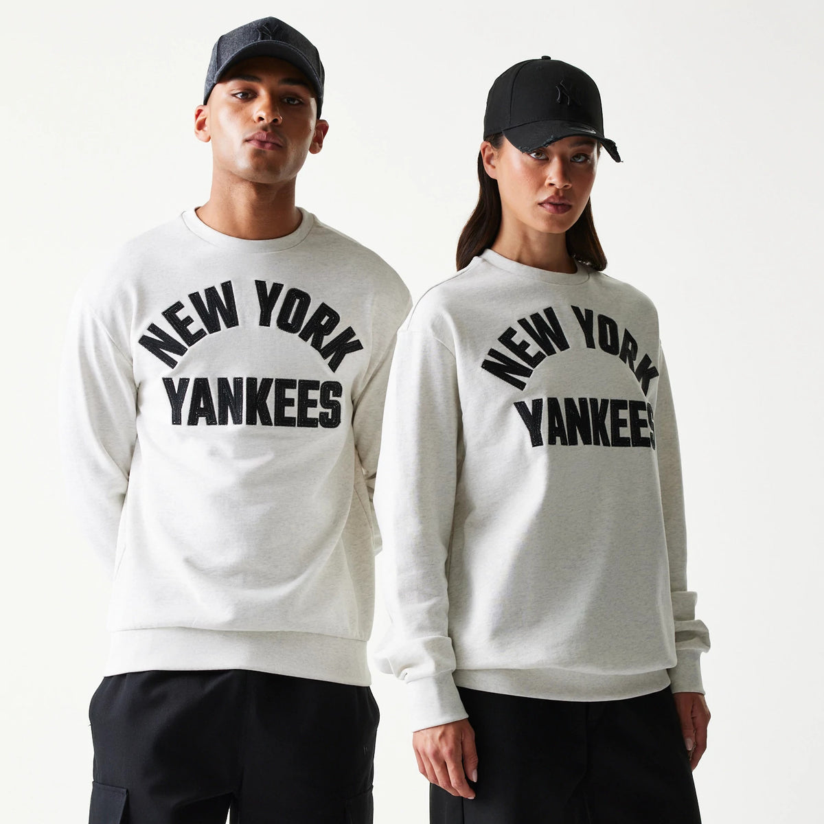 New Era New York Yankees MLB Graphic Pastel Grey Oversized Crew Neck Sweater, front view on models with matching black caps, featuring black New York Yankees lettering across the chest and ribbed cuffs at the sleeves.