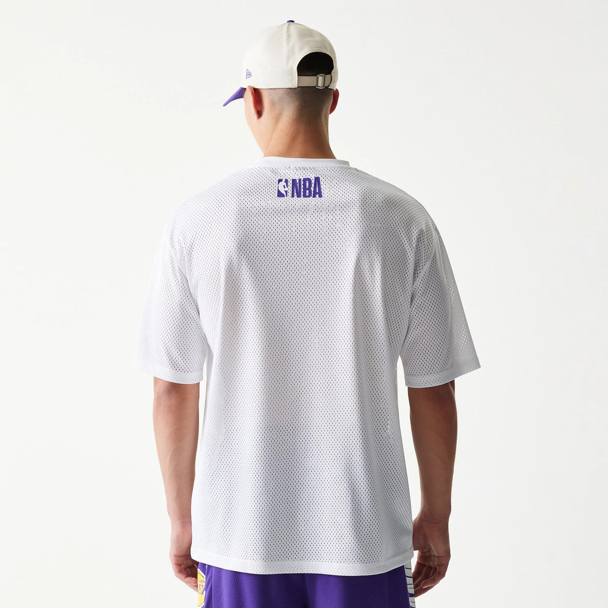New Era LA Lakers NBA Graphic Mesh White T-Shirt, back view on a model wearing purple shorts and a white and purple cap, showing a blue printed NBA logo across the upper back of the white mesh fabric.