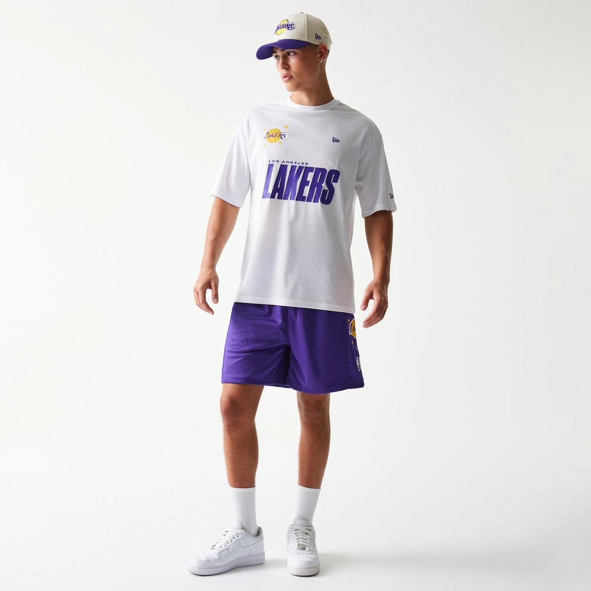 New Era LA Lakers NBA Graphic Mesh White T-Shirt, front view on-model showing a large purple Lakers printed logo across the chest, a small raised Lakers emblem on the upper left, and paired with purple shorts and a matching cap.
