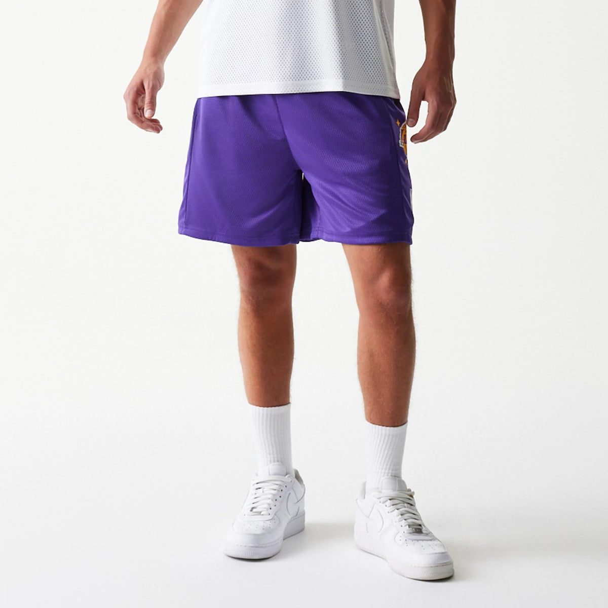 New Era LA Lakers NBA Mesh Purple Shorts, front view on-model showing the purple colour, featuring an embroidered LA Lakers logo on the side near the hem, paired with white socks and trainers.