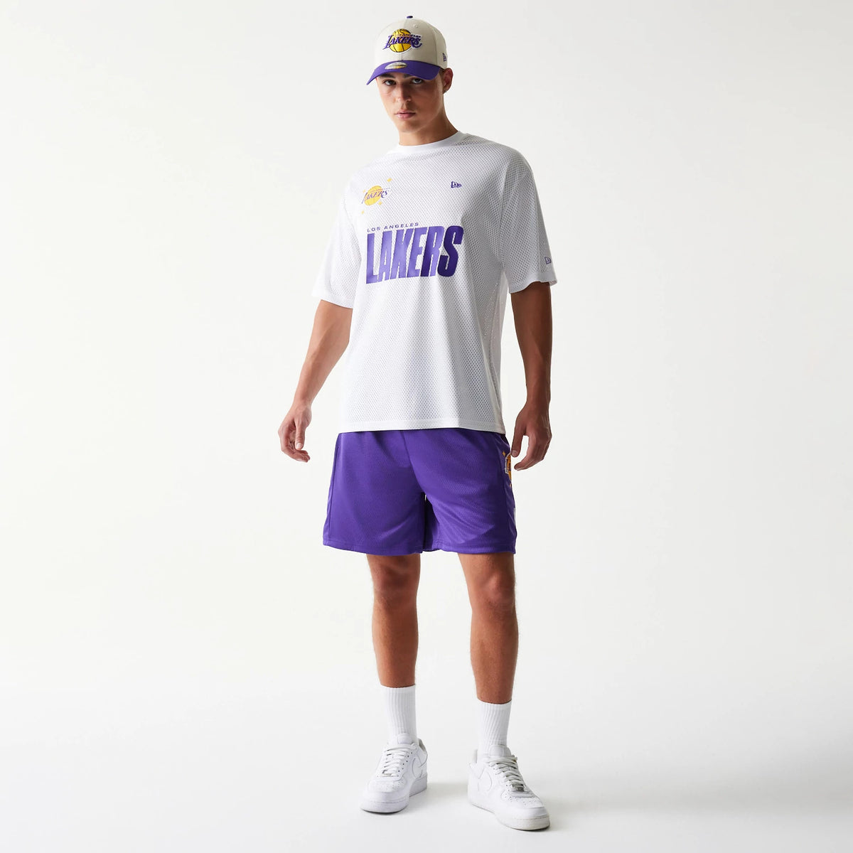 New Era LA Lakers NBA Mesh Purple Shorts, front view on-model with a white Lakers T-shirt and cap, showing the rich purple colour of the shorts with the Lakers logo prominently displayed on the left leg.