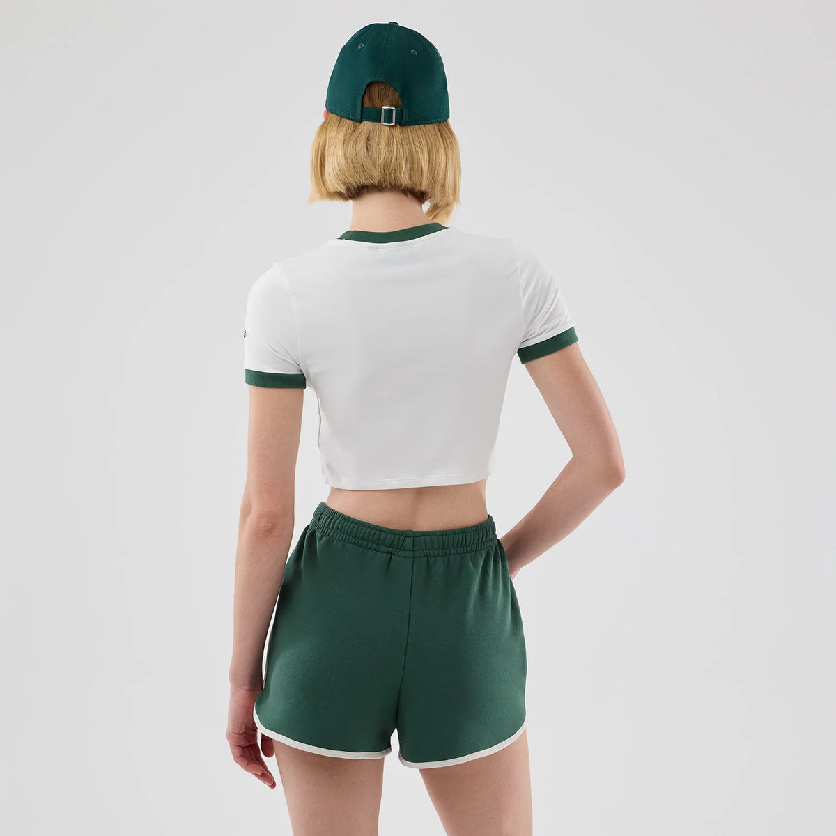 Womens New Era White Cropped Slim T-shirt, back view on a model wearing matching green shorts and cap, showing green ribbed trim around the collar and sleeves with a small embroidered New Era logo on the left sleeve.