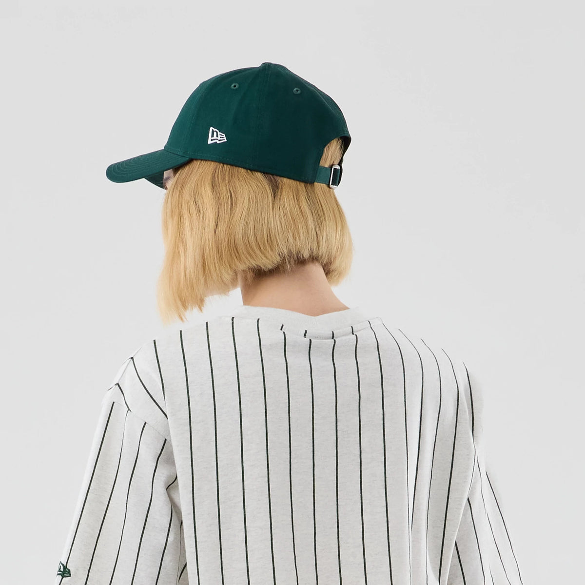 Womens New Era Pinstripe Pastel Grey Cropped T-shirt, back view on a model wearing a dark green cap, showing thin vertical black stripes across the light grey fabric and the New Era logo embroidered on the left sleeve.