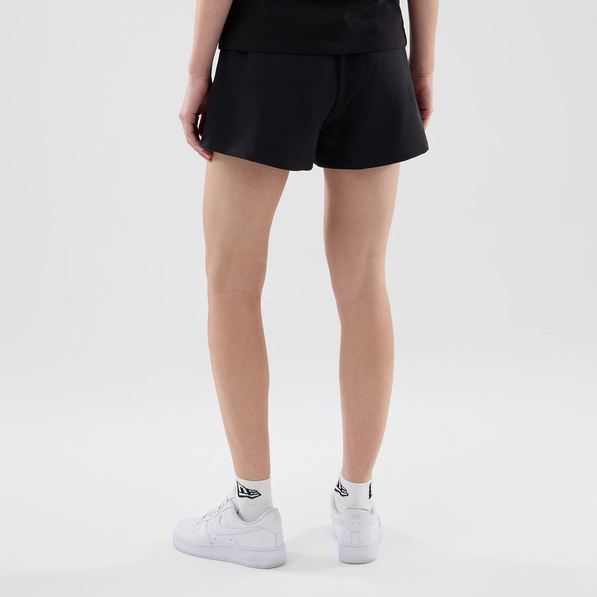 New Era New York Yankees Womens MLB Infill Black Shorts, back view on-model showing the plain black fabric with a relaxed fit at the hem, paired with white trainers and socks featuring the New Era logo on the back.