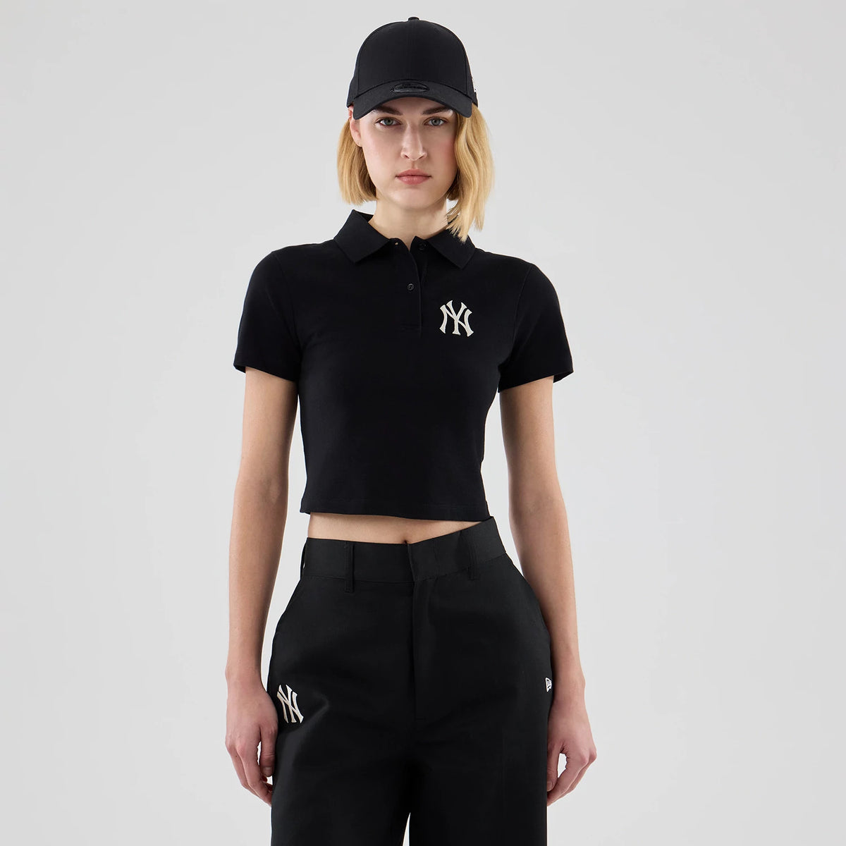 New Era New York Yankees Womens MLB Black Cropped Slim Polo T-shirt, front view showing the embroidered Yankees logo on the left chest, on-model styled with matching black trousers and cap.