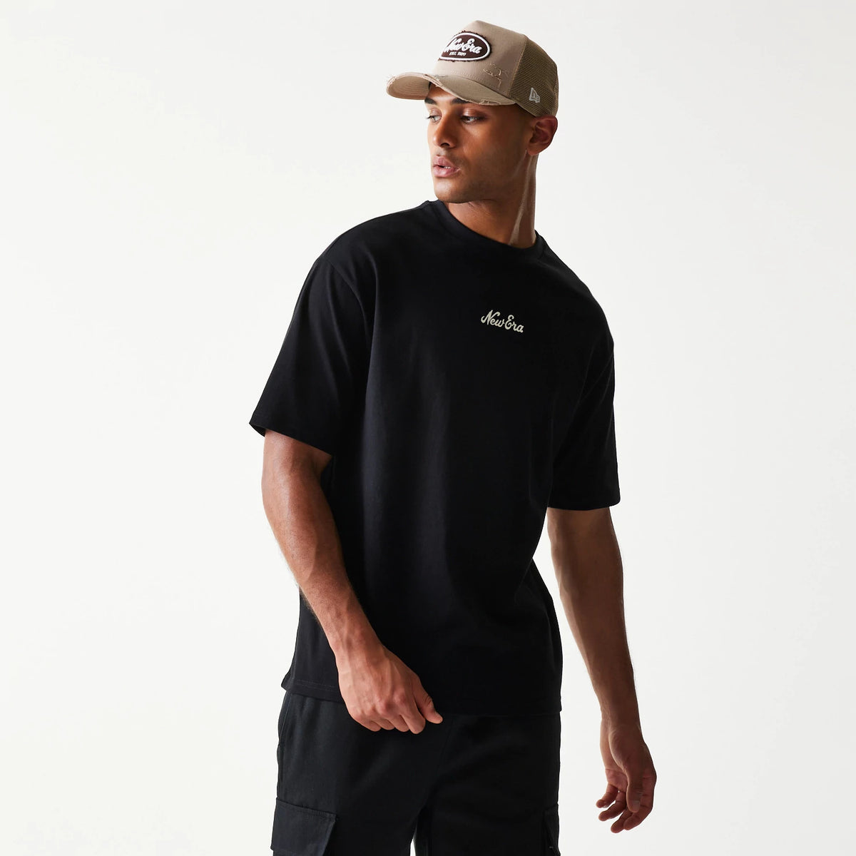 New Era Heritage Script Black T-Shirt, front view on a model paired with dark trousers, featuring an embroidered script logo in white on the left chest area, the simple black design accentuating the casual look.