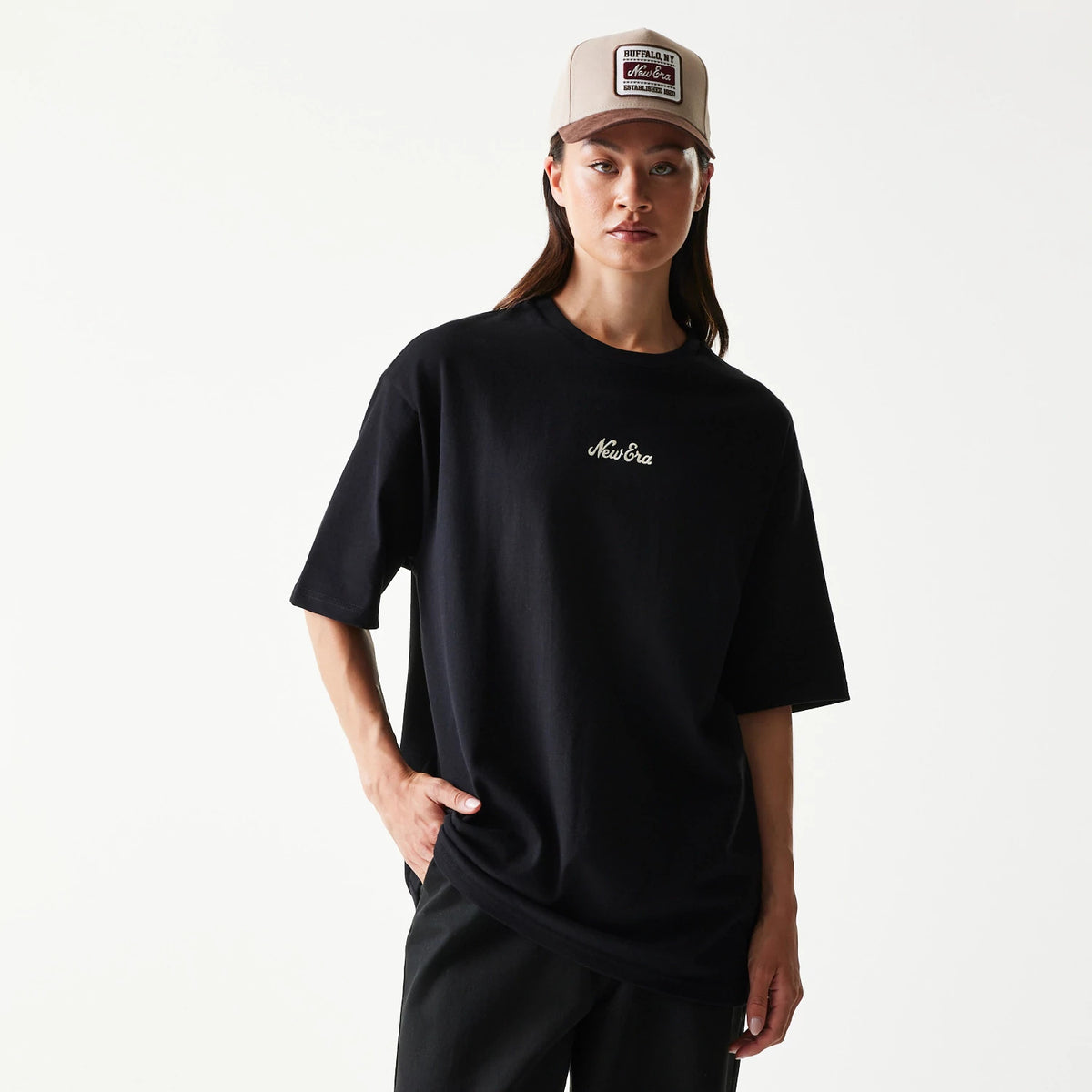 New Era Heritage Script Black T-Shirt, front view on a model showing a relaxed black silhouette with a white embroidered New Era logo centred on the chest, styled with a neutral cap.