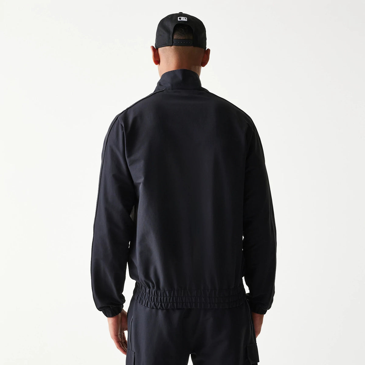 New Era New York Yankees MLB Black Track Jacket, back view on-model displaying the black back panel and an elasticated waist, paired with a black cap featuring an embroidered logo on the back.
