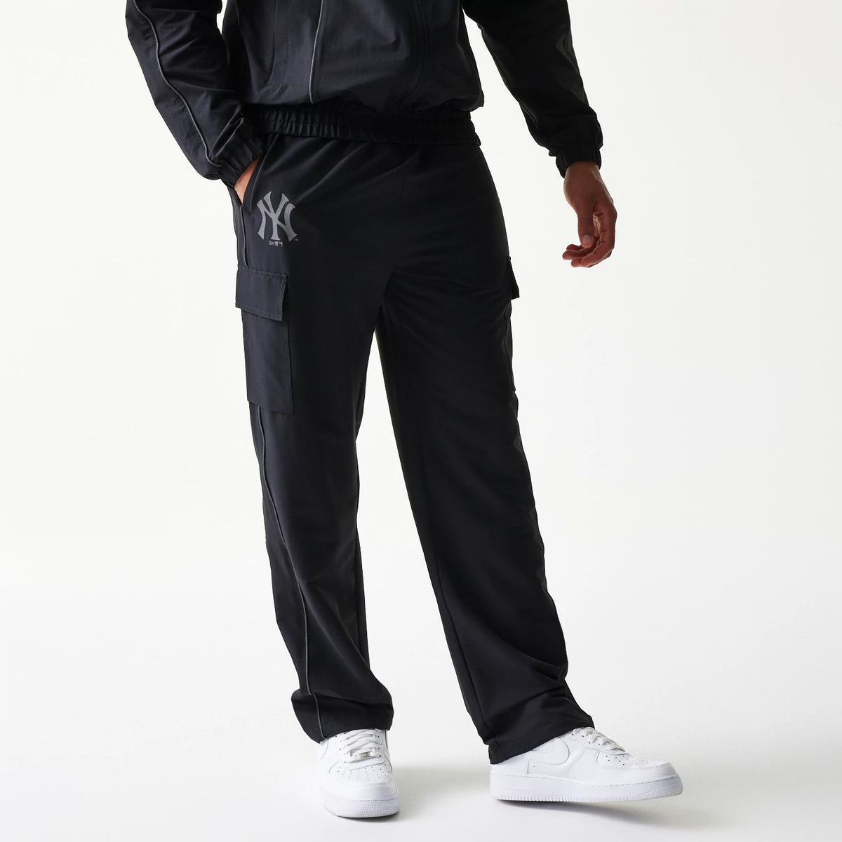 New Era New York Yankees MLB Utility Black Straight Leg Cargo Joggers, front view on a model wearing white trainers and a matching jacket, with the grey Yankees logo on the left thigh and cargo pockets on each side.