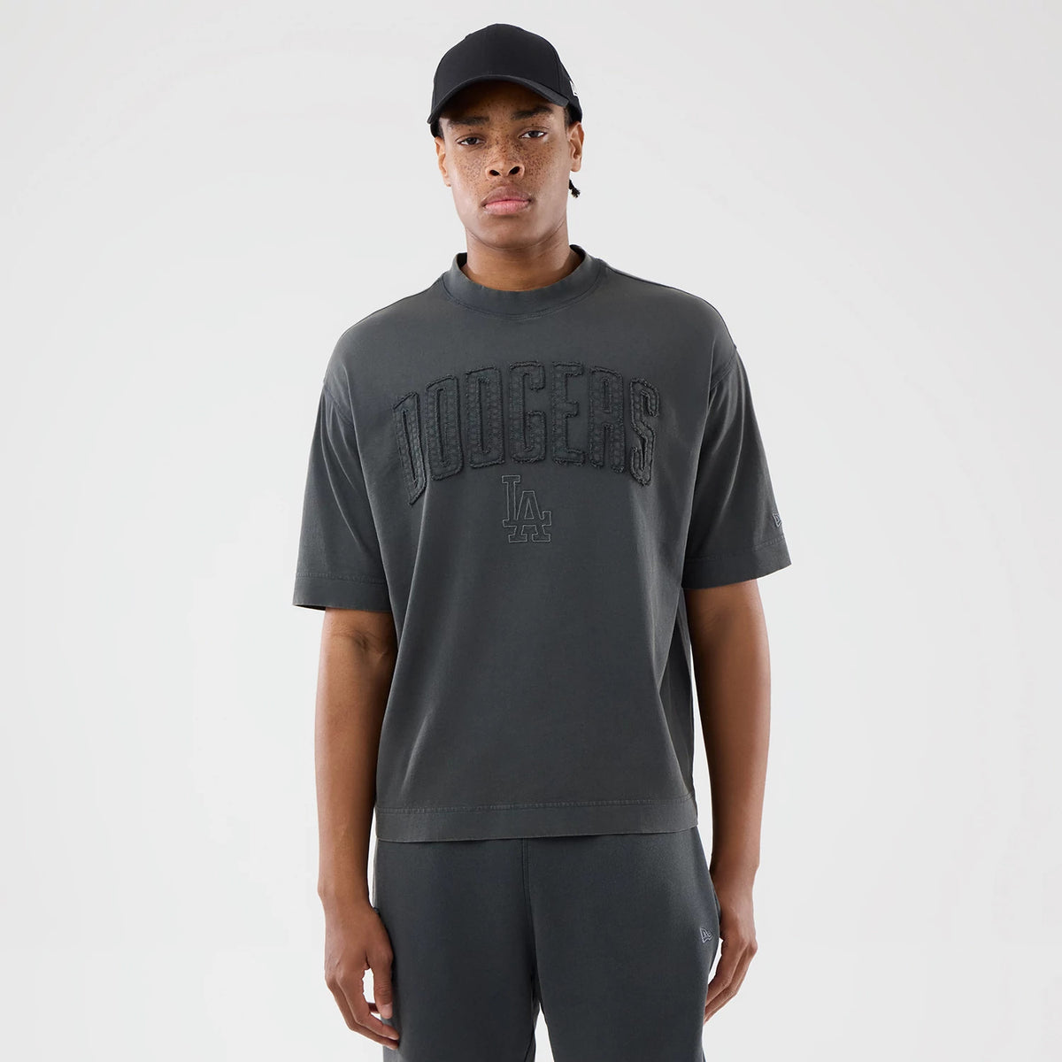 New Era LA Dodgers MLB Washed Dark Grey T-Shirt, front view on a model showcasing large embroidered Dodgers lettering across the chest with a small LA logo underneath, the dark grey fabric complementing a black cap worn by the model.
