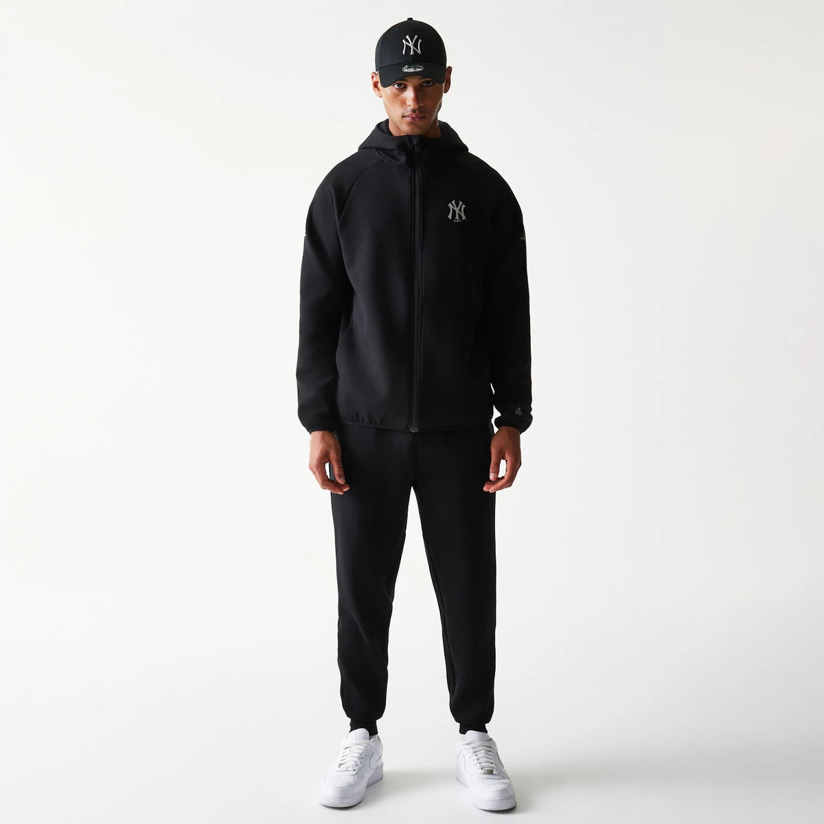 New Era New York Yankees MLB Black Full Zip Hoodie, front view on-model showing the hooded black jacket with an embroidered Yankees logo on the chest, styled with matching black trousers and a black cap adorned with the Yankees logo.