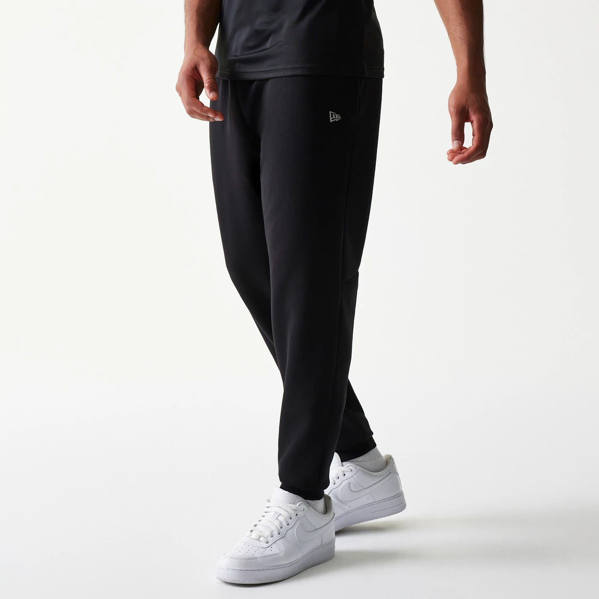 New Era New York Yankees MLB Black Joggers, on-model front view showing the New Era flag logo embroidered on the left thigh of the tapered black joggers, paired with white trainers and a black t-shirt.