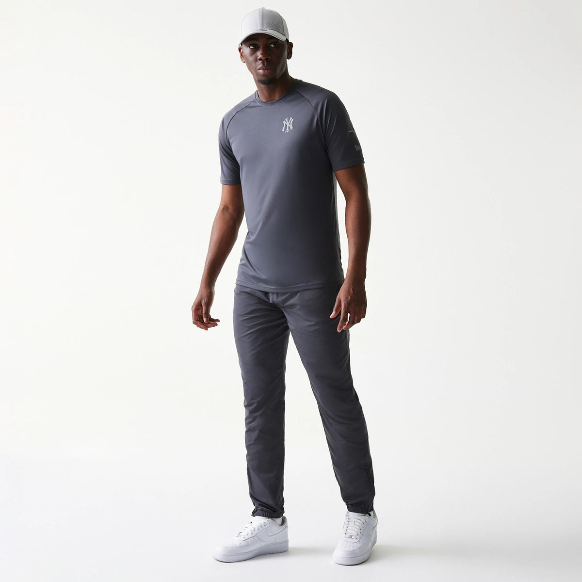 New Era New York Yankees MLB Dark Grey Track Pants, front view on a model with a grey tee and cap, showcasing a slim fit and elasticated ankles for a look, paired with white trainers that accentuate the outfit.