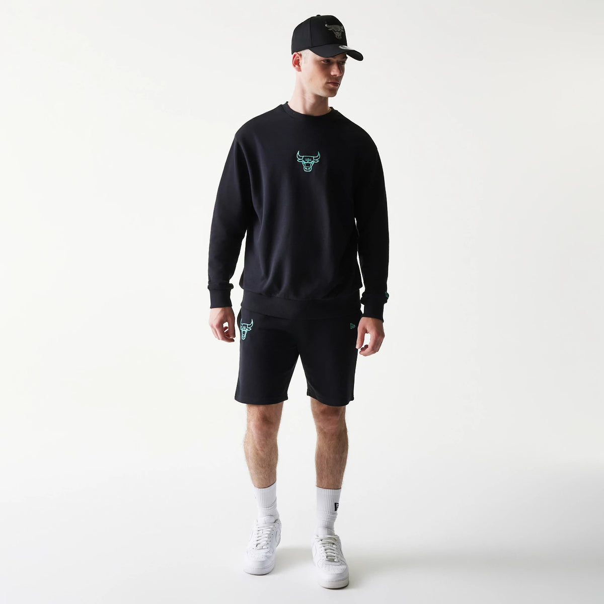 New Era Chicago Bulls NBA Black Oversized Crew Neck Sweater, front view on a model with matching black shorts and cap, showing the turquoise embroidered Bulls logo on the chest and left leg, with relaxed fit and ribbed cuffs.