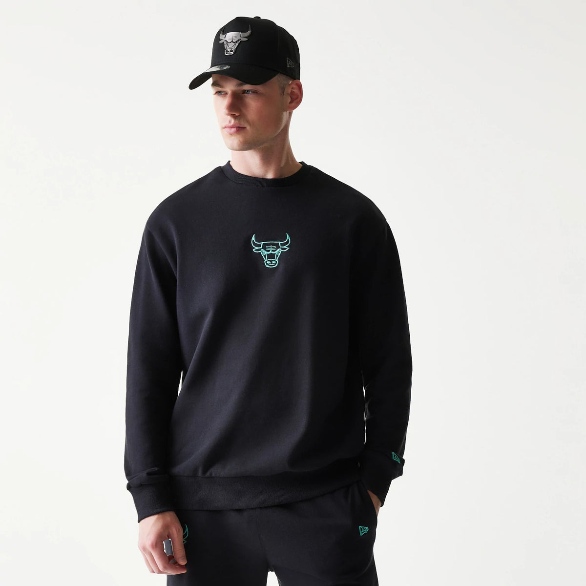 New Era Chicago Bulls NBA Black Oversized Crew Neck Sweater, front view on a model with matching black trousers and cap, featuring the mint green embroidered Bulls logo on the chest and the New Era flag logo on the left cuff.