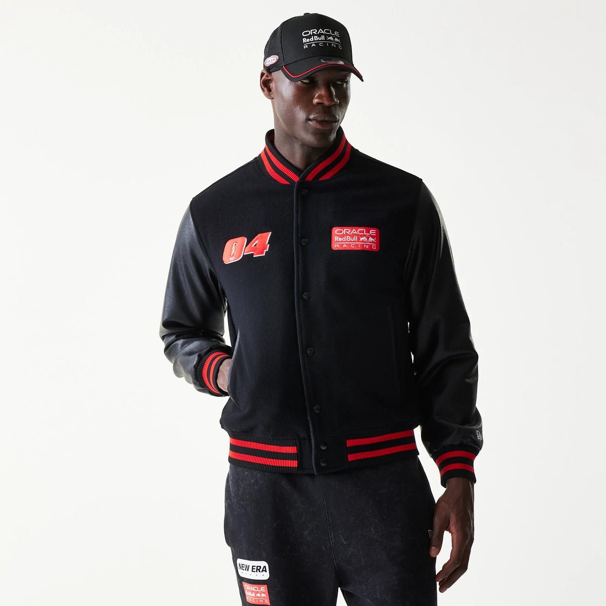 New Era Red Bull Racing Black Varsity Jacket, front view on-model showing an embroidered number 04 on the left chest, a Red Bull Racing patch on the right chest, and red and black striped cuffs and hem with a snap button closure.