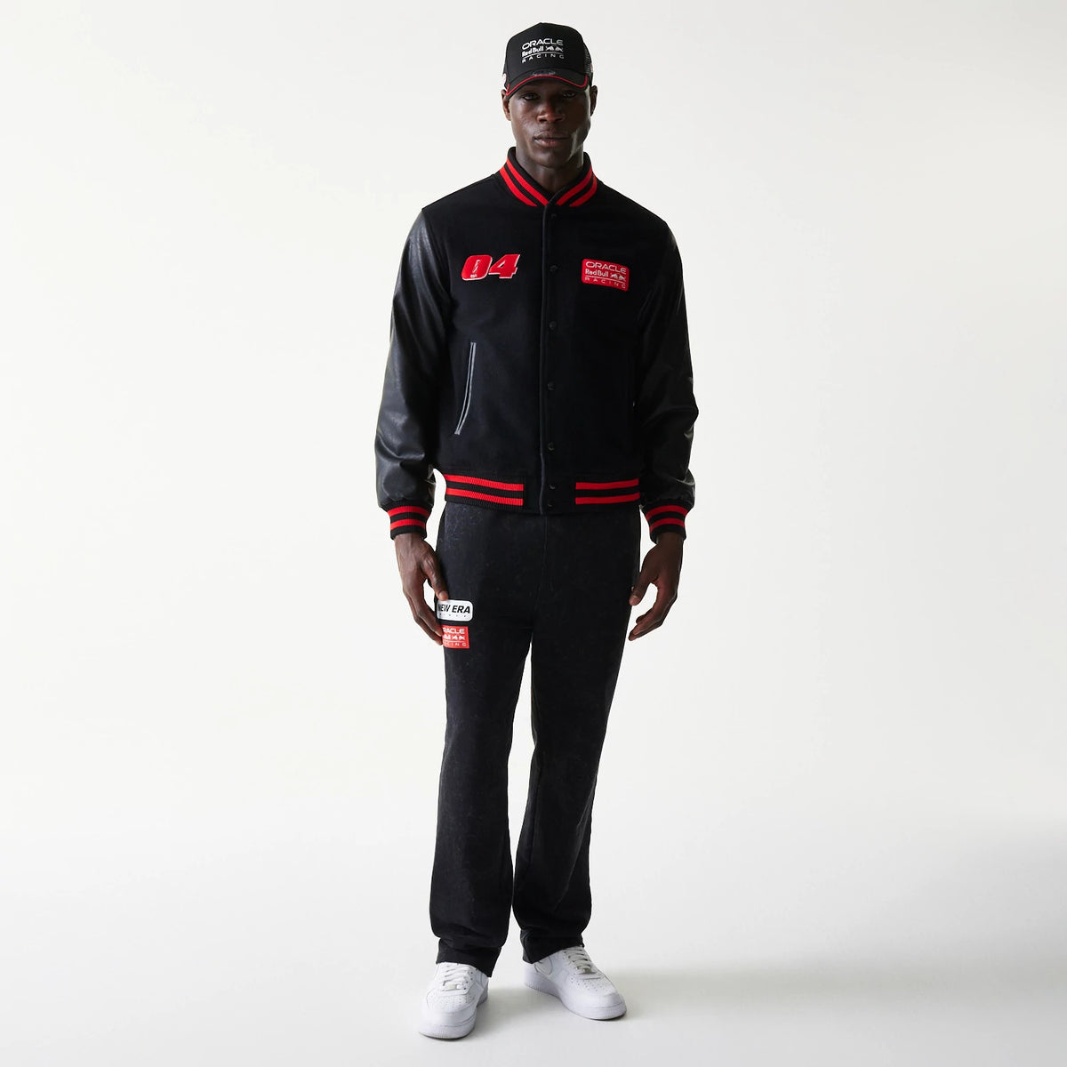 New Era Red Bull Racing Black Varsity Jacket, front view on-model displaying red embroidered 04 on the right chest, a red patch on the left chest, striped red and black cuffs and hem, matched with a black cap and trousers.