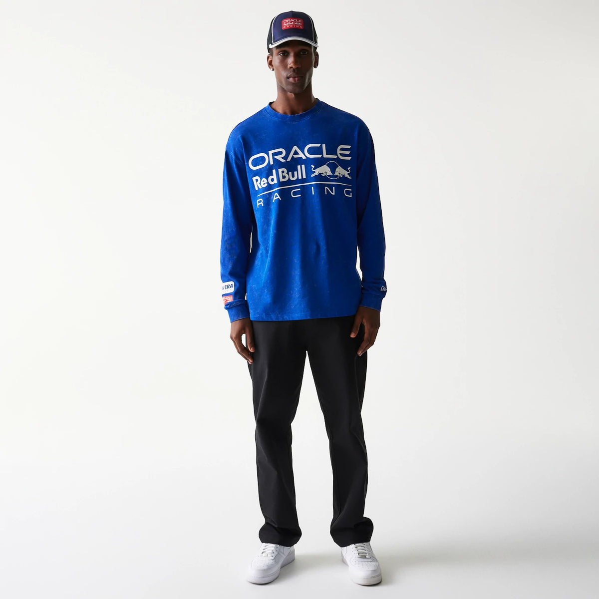 New Era Red Bull Racing Linear Graphic Blue Long Sleeve T-Shirt, front view on-model showing a prominent white printed Oracle Red Bull Racing logo across the chest and smaller logos on the sleeves, paired with black trousers and white trainers.