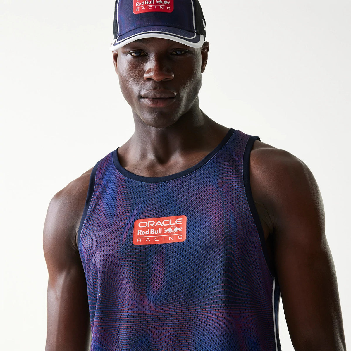 New Era Red Bull Racing Dark Blue Mesh Tank Top, on-model front view featuring an orange Red Bull Racing logo on the centre chest, with the dark blue mesh fabric showing purple accents and a scoop neckline.