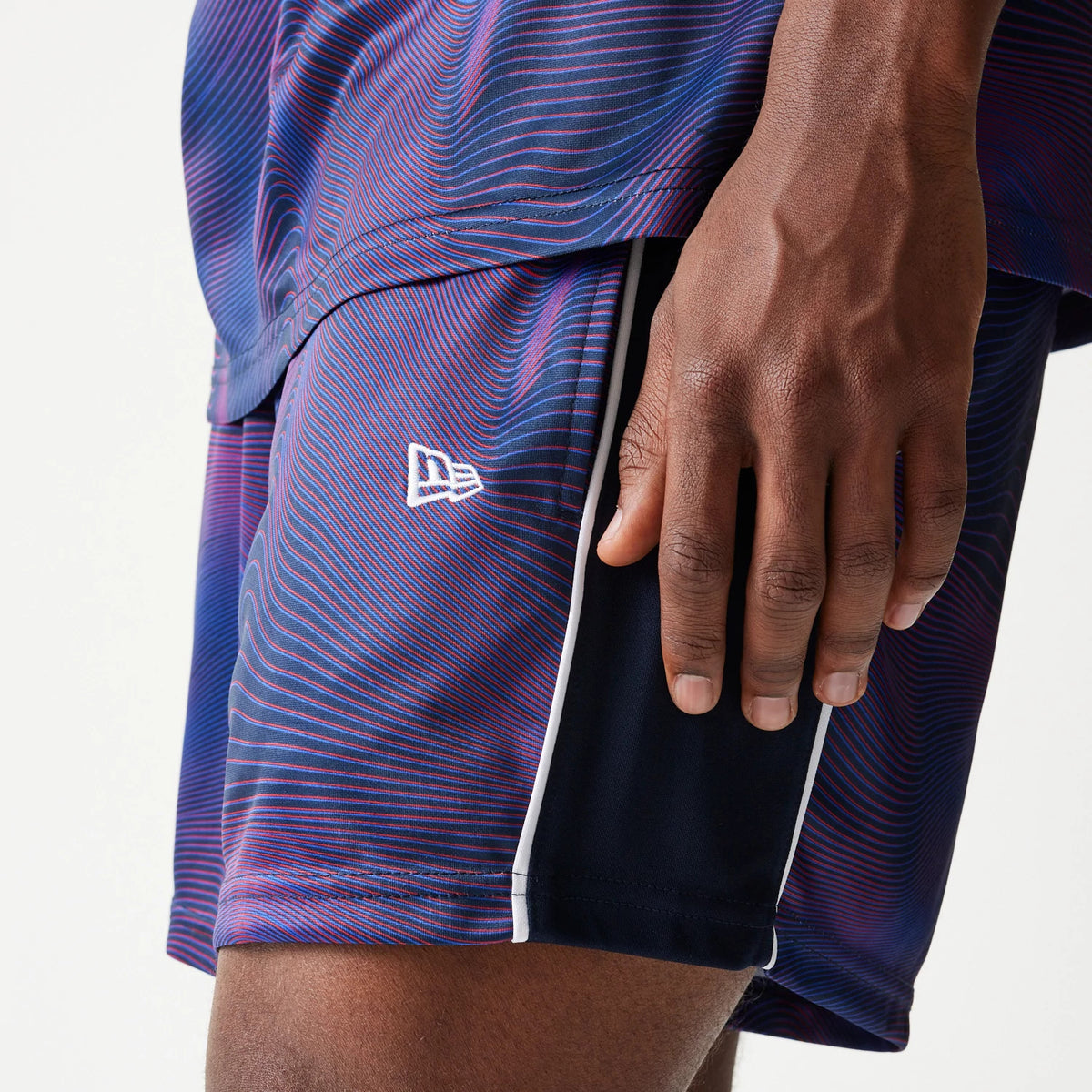 New Era Red Bull Racing All Over Print Dark Blue Football Shorts, side view showing the swirling blue and red pattern with white embroidered New Era logo near the hem, and vertical white piping along the edges.