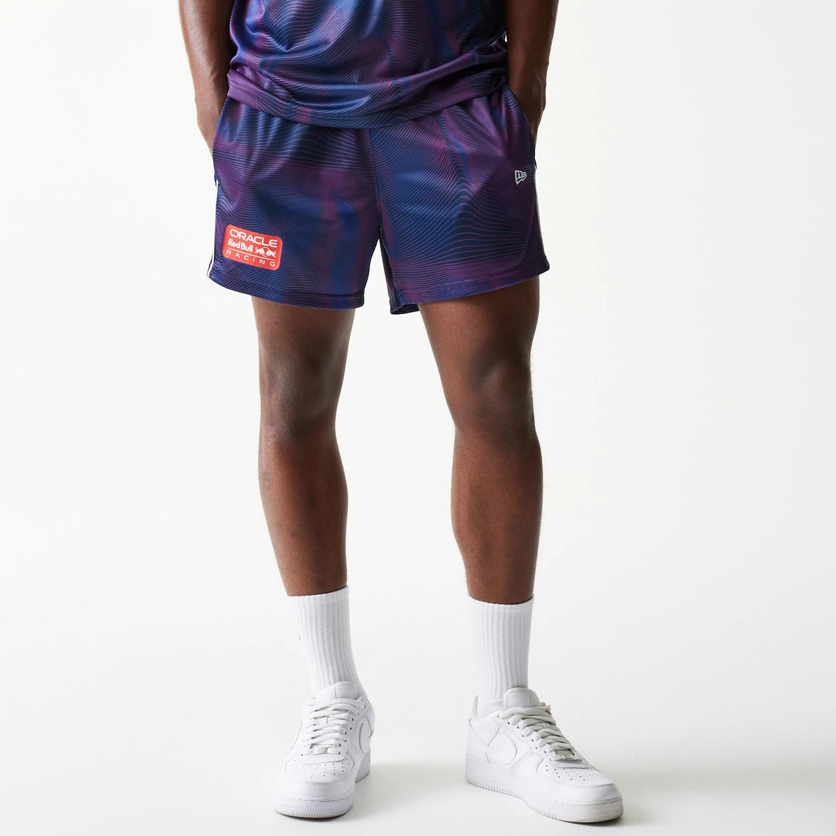 New Era Red Bull Racing All Over Print Dark Blue Football Shorts, front view on a model with white trainers showing a wave pattern, an embroidered New Era flag above the hem, and a red and white Red Bull Racing patch on the left leg.