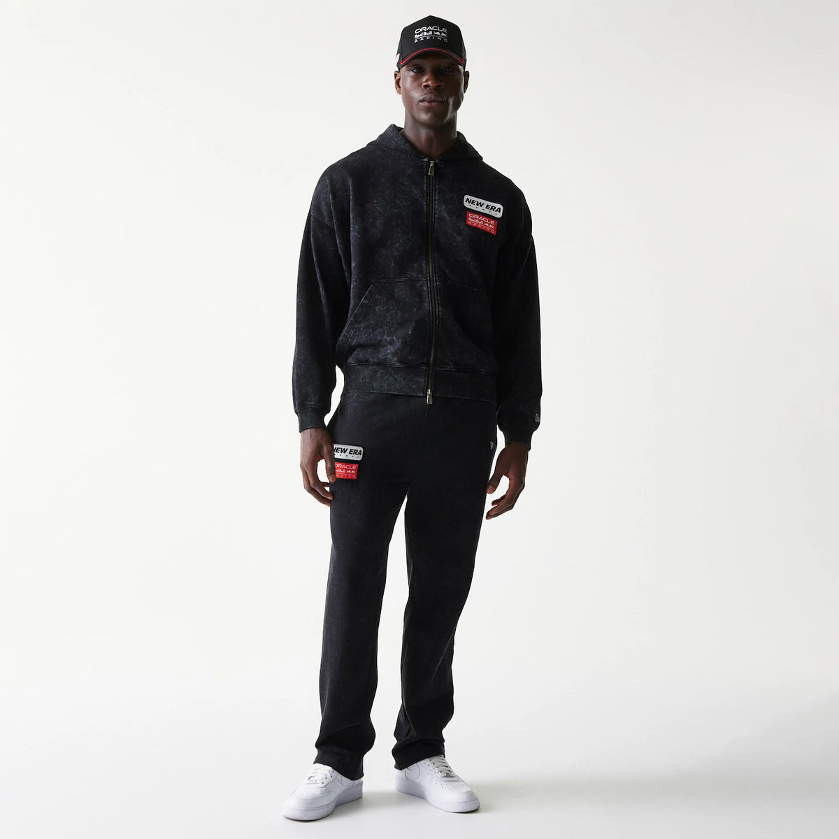 New Era Red Bull Racing Straight Leg Black Straight Leg Joggers, front view on a model with matching black hoodie and cap, featuring a New Era and Red Bull patch on the left leg above the knee and a relaxed fit over the shoes.
