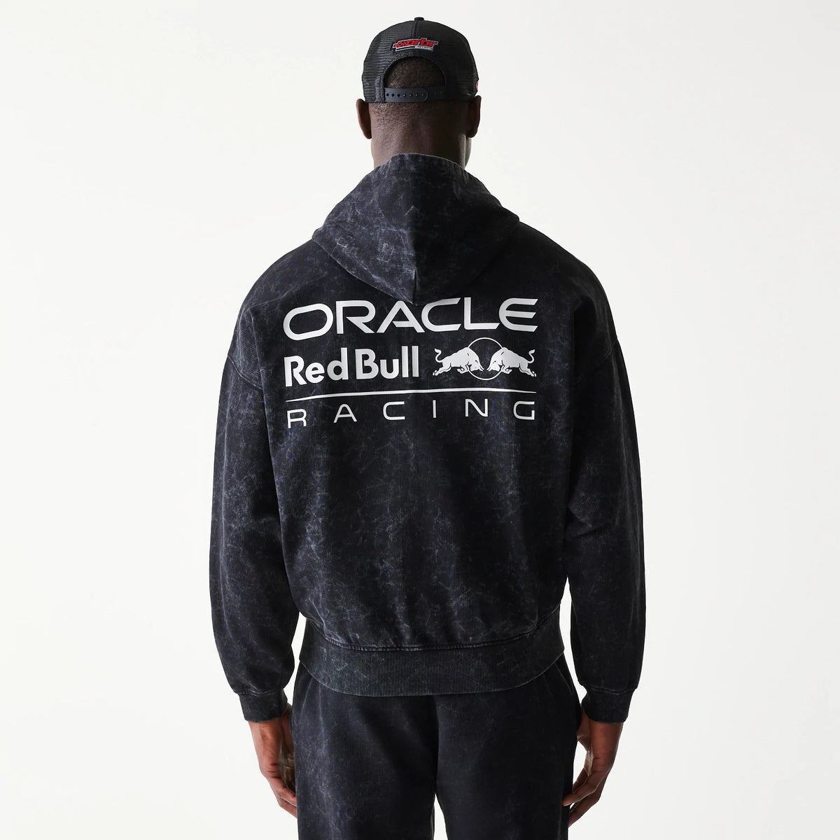 New Era Red Bull Racing Dropped Shoulder Black Dropped Shoulder Full Zip Hoodie, back view on a model styling matching trousers and cap, featuring large printed Oracle Red Bull Racing lettering across the upper back under the hood.