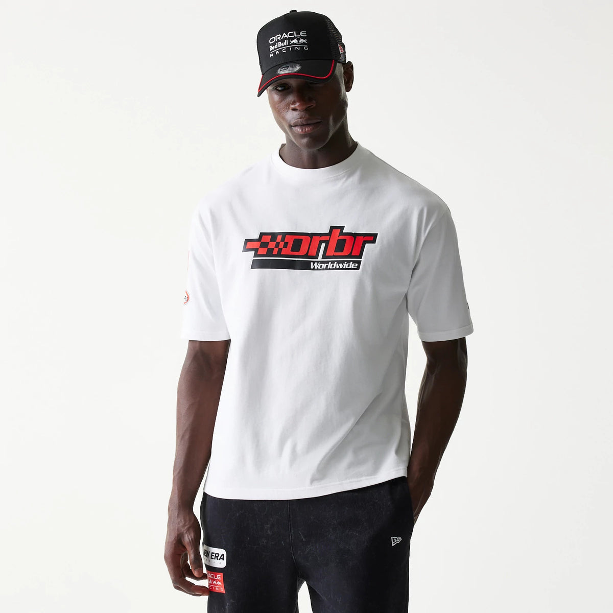 New Era Red Bull Racing Graphic White Oversized T-Shirt, front view on a model featuring red and black racing graphics and text across the chest, styled with a black cap and dark trousers showing New Era branding.