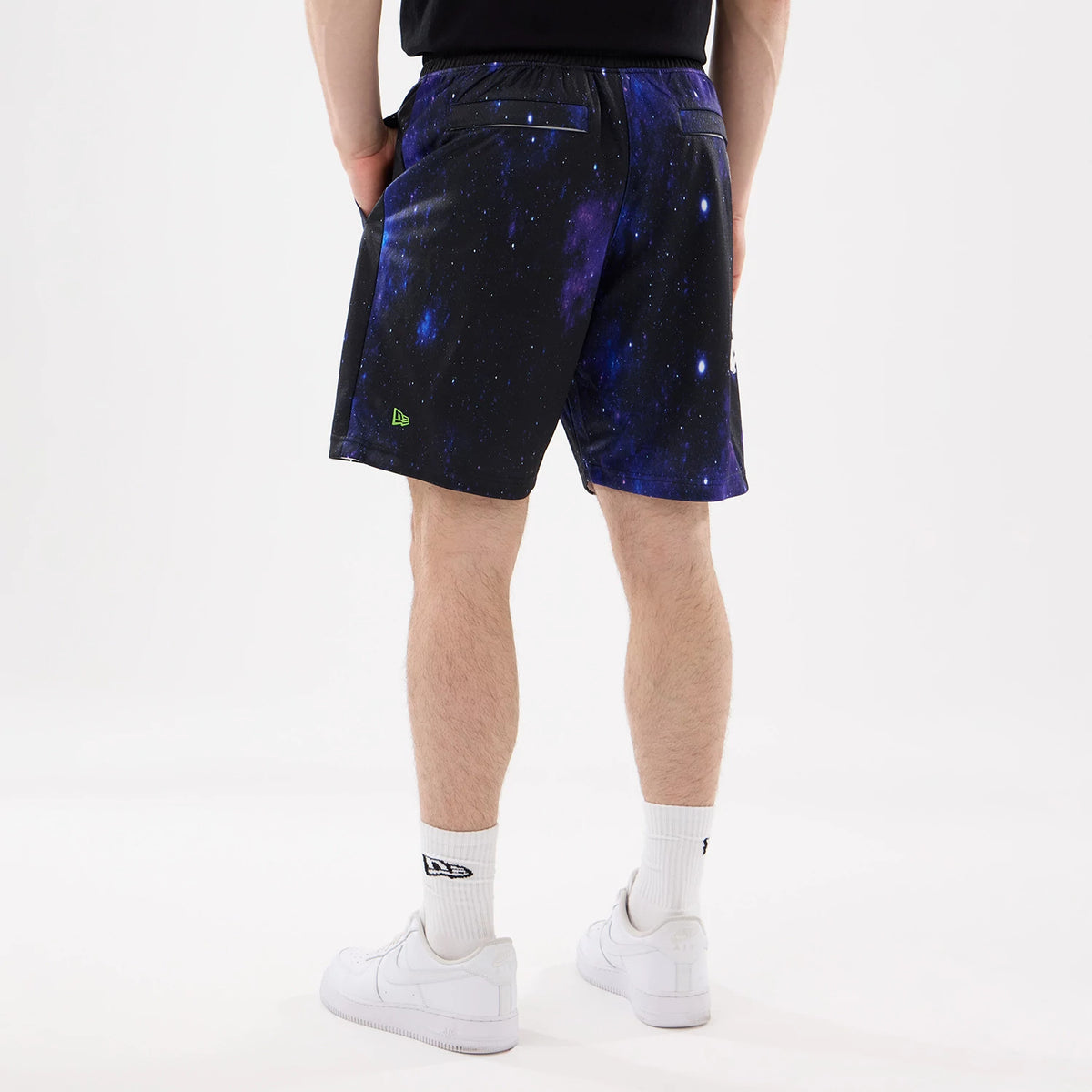 New Era Space Jam Warner Brothers Space Jam White Oversized Shorts, back view on-model showing a galaxy print design with black waistband and green embroidered New Era logo on the lower left, model styled with white trainers and socks.
