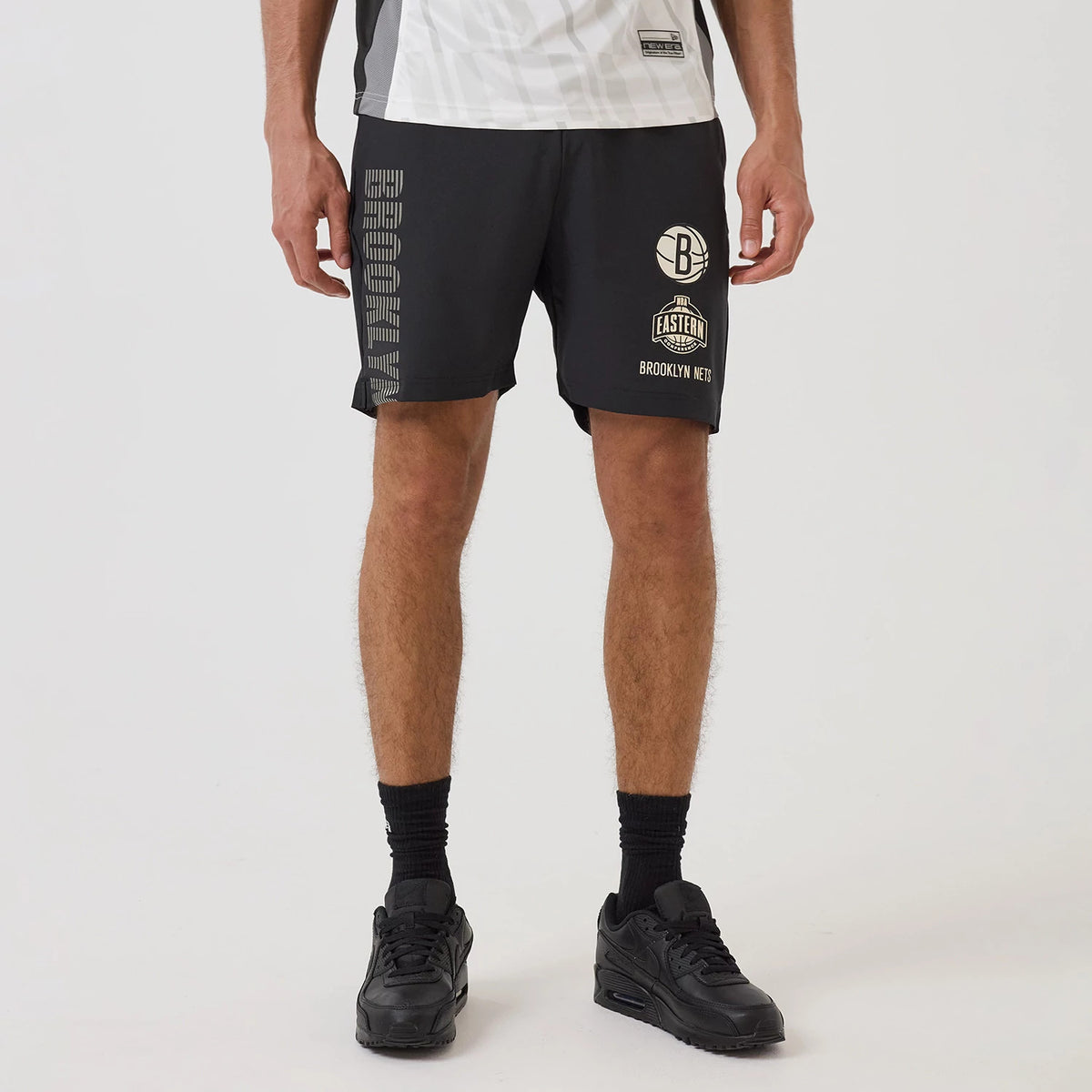 New Era Brooklyn Nets NBA Match Black Oversized Shorts, front view showing black shorts with Brooklyn Nets and NBA Eastern logos printed on the right leg and vertical BROOKLYN lettering printed along the left leg, styled with black trainers.