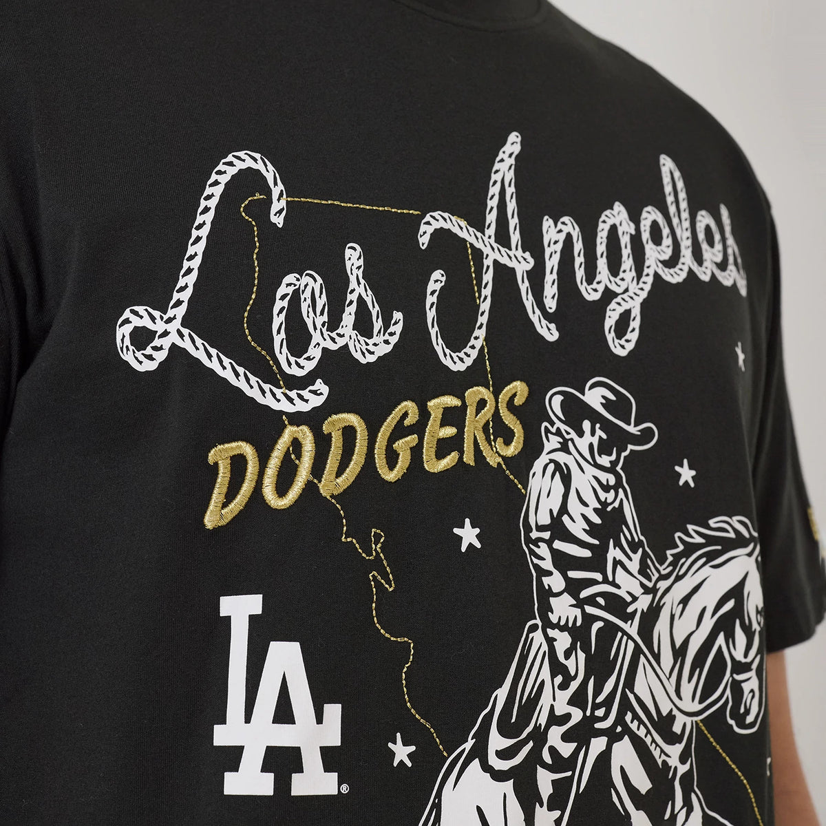 New Era LA Dodgers MLB Cowboy Graphic Black T-Shirt, front view showing white embroidered Los Angeles and gold Dodgers lettering across the chest, a printed cowboy illustration, and the white LA logo at the bottom.